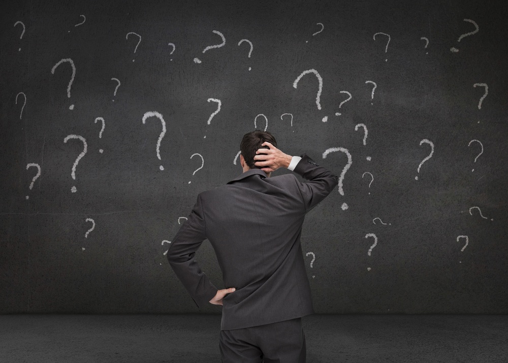 Rear view of a doubtful businessman looking at various question marks drawn on a grey wall